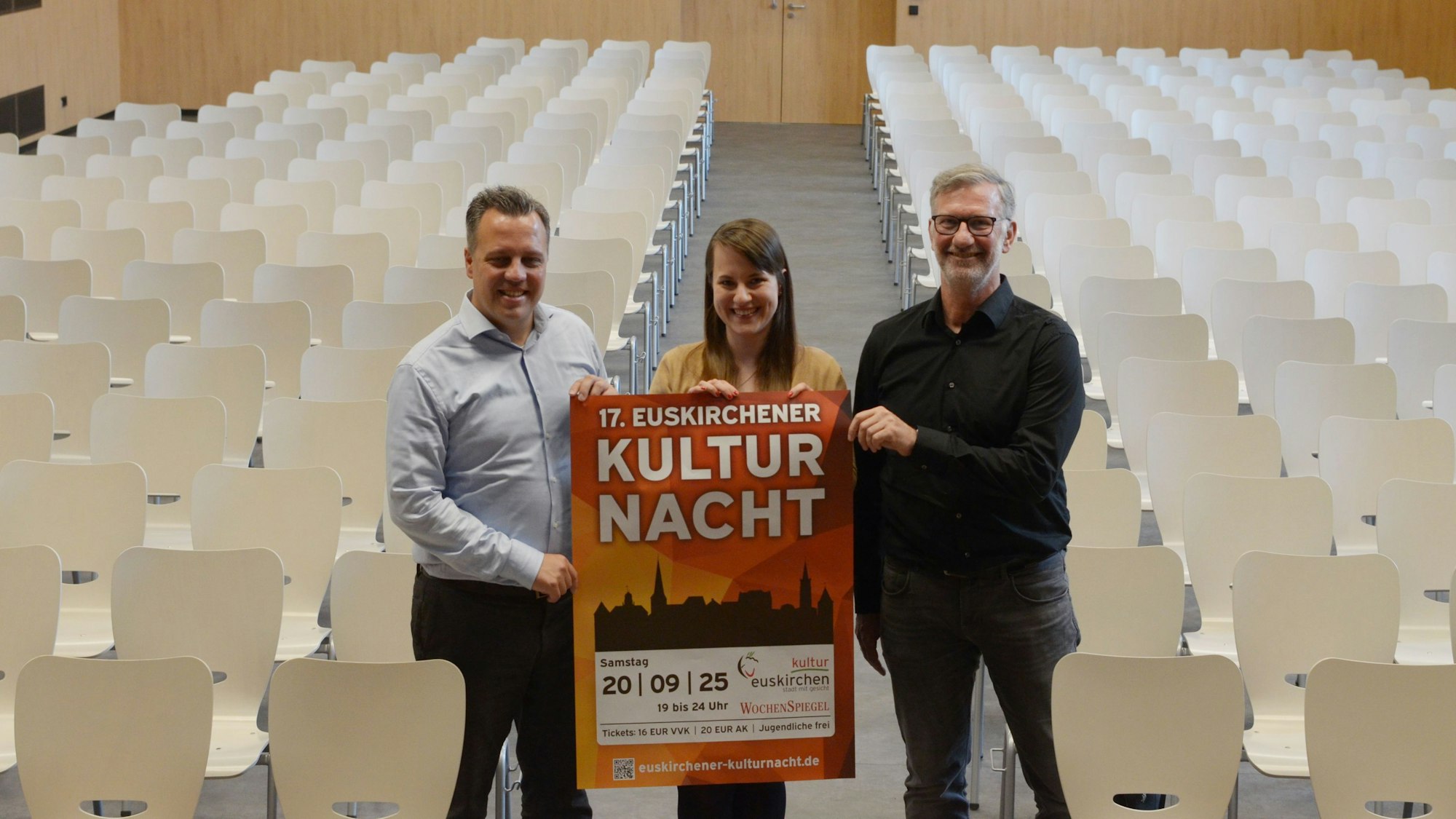 Sacha Reichelt, Nina Kaiser-Havertz und Thomas Müller halten ein Plakat auf dem „17. Euskirchener Kultur Nacht“ steht in einer bestuhlten Aula in die Kamera.
