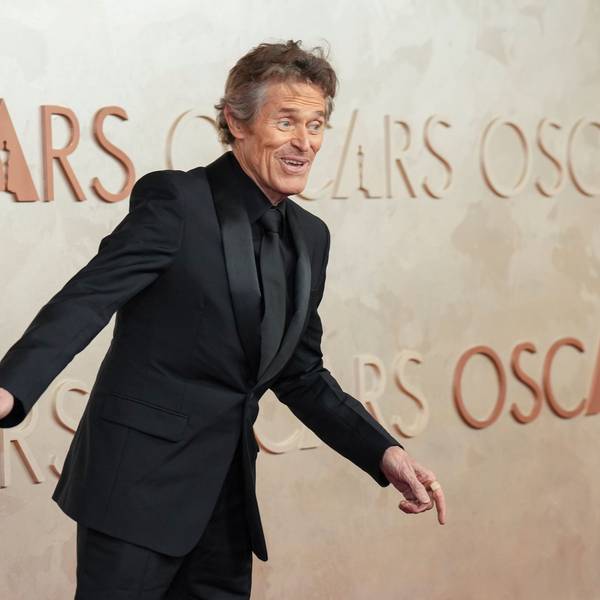 Willem Dafoe, im schwarzen Anzug mit schwarzer Krawatte, gestikuliert für die Fotografen.