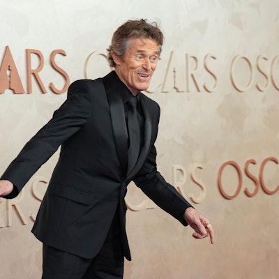 Willem Dafoe, im schwarzen Anzug mit schwarzer Krawatte, gestikuliert für die Fotografen.