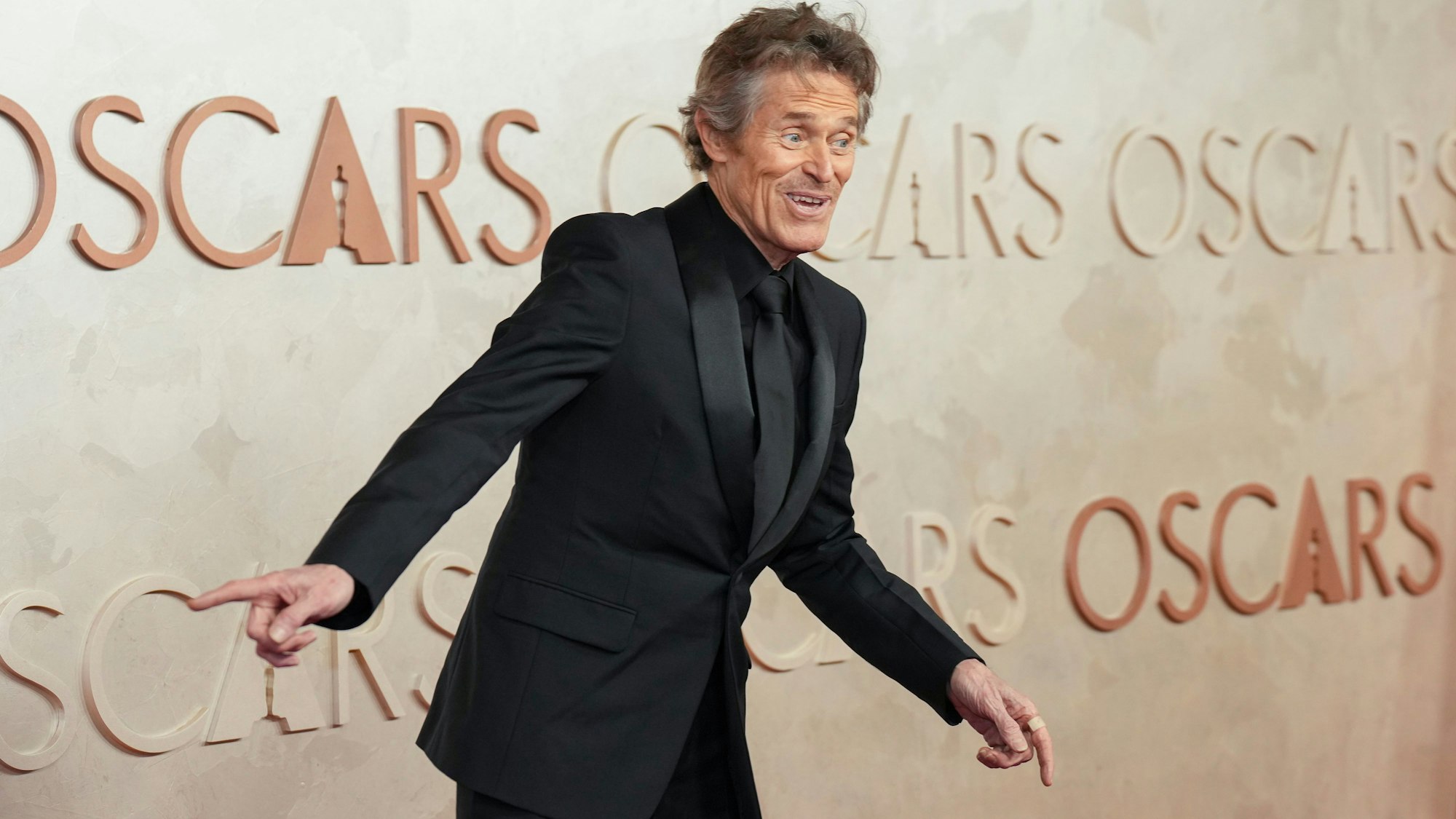 Willem Dafoe, im schwarzen Anzug mit schwarzer Krawatte, gestikuliert für die Fotografen.