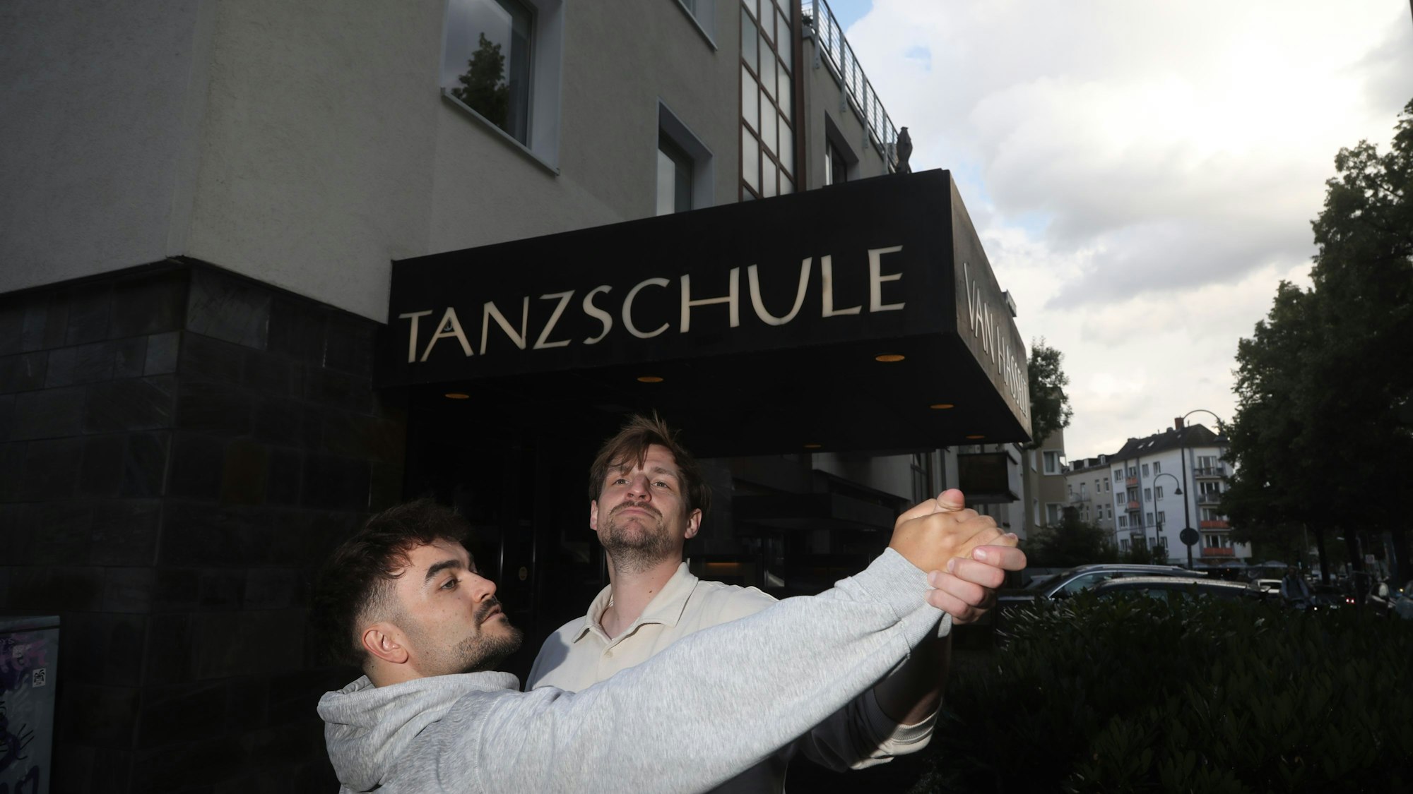 Zwei junge Männer tanzen miteinander auf offener Straße vor dem Eingang einer Tanzschule.