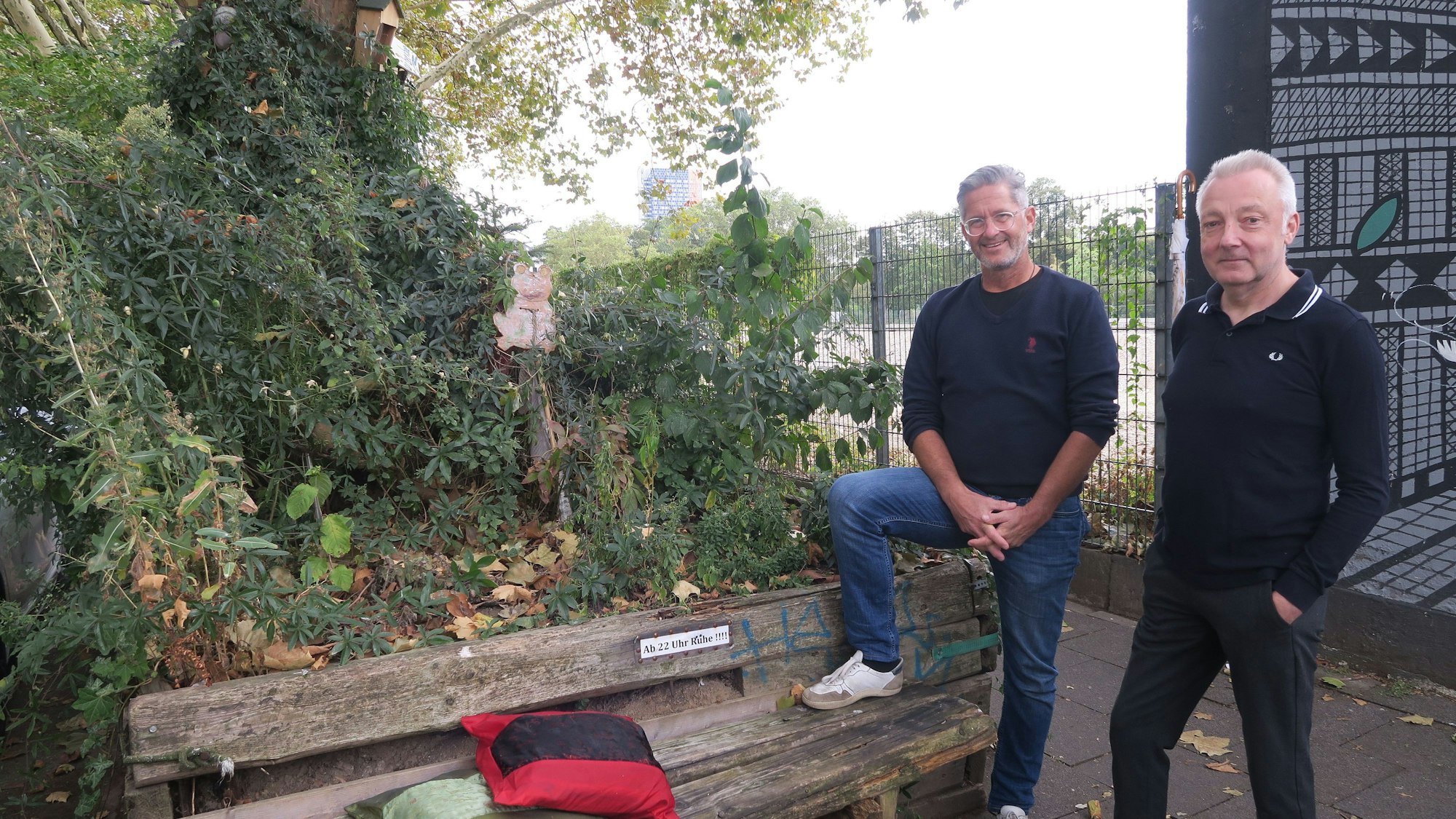 Zwei Männer stehen neben einer Bank auf einem Bürgersteig. Hinter der Bank steht ein alter Baum.