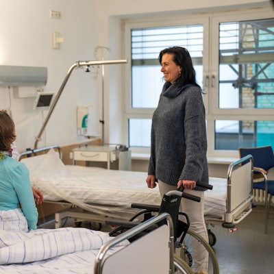 Eine Frau sitzt auf einem Krankenhausbett, rechts daneben steht eine Frau mit Rollstuhl und rechts daneben steht eine Frau, die der Sitzenden die Hand auf den Rücken legt.