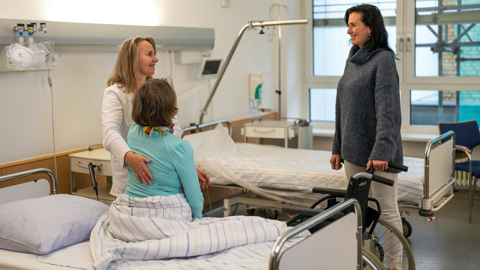 Eine Frau sitzt auf einem Krankenhausbett, rechts daneben steht eine Frau mit Rollstuhl und rechts daneben steht eine Frau, die der Sitzenden die Hand auf den Rücken legt.