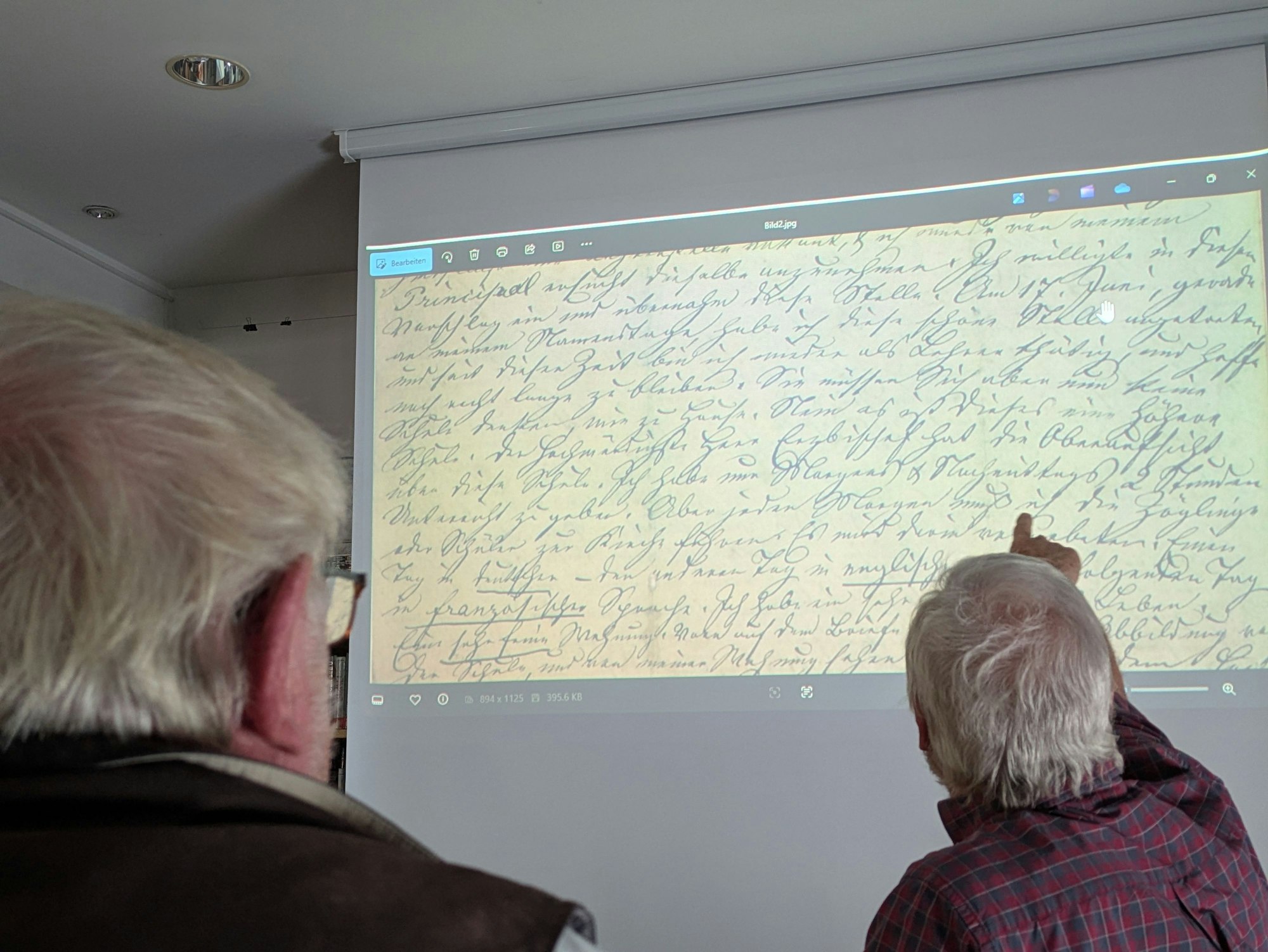 Zwei Männer sitzen vor einer Leinwand, auf die ein Brief in Kurrentschrift geworfen ist.