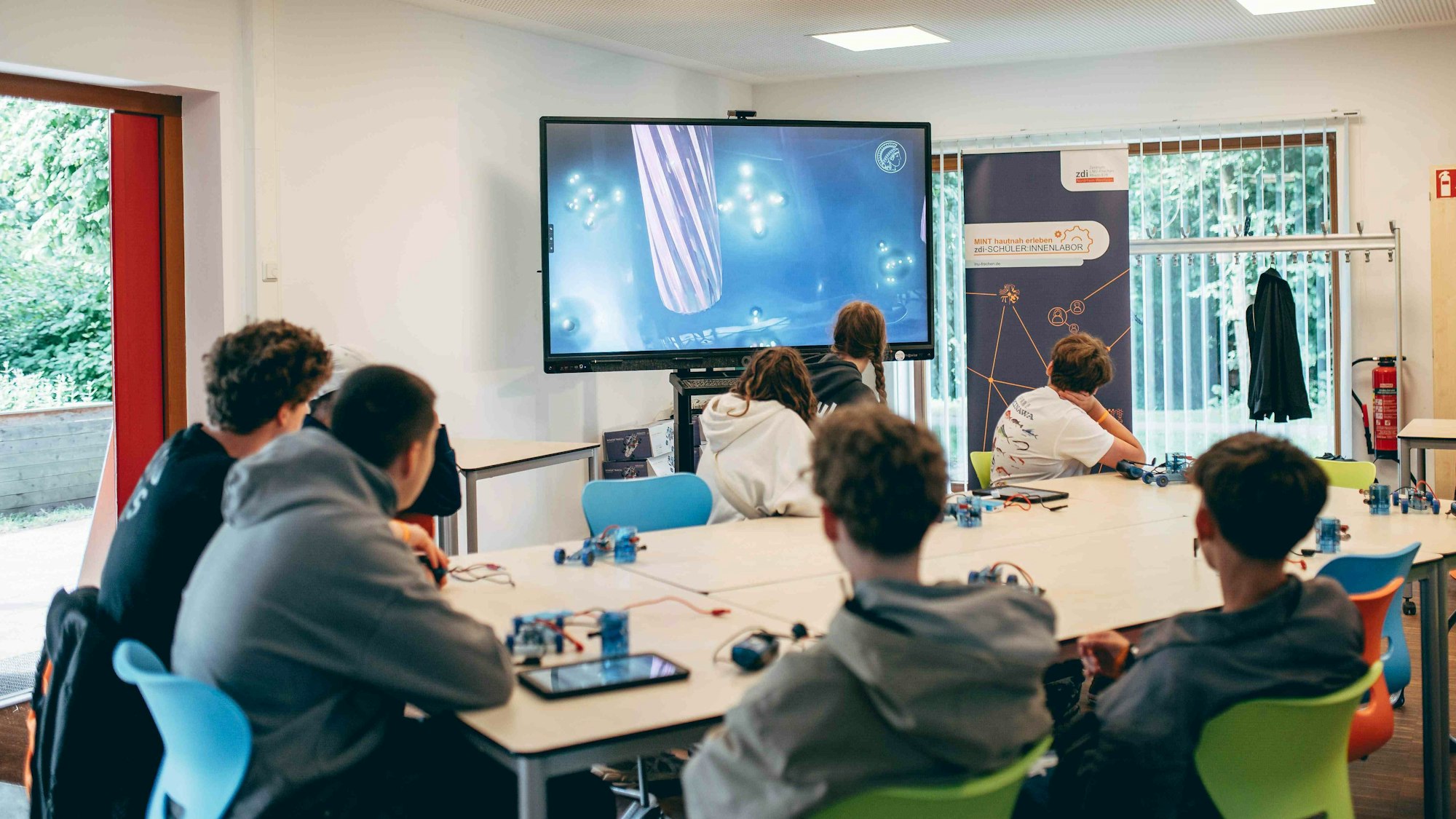 Schülerinnen und Schüler sitzen an einem Tisch und schauen einen Film auf einem Bildschirm.