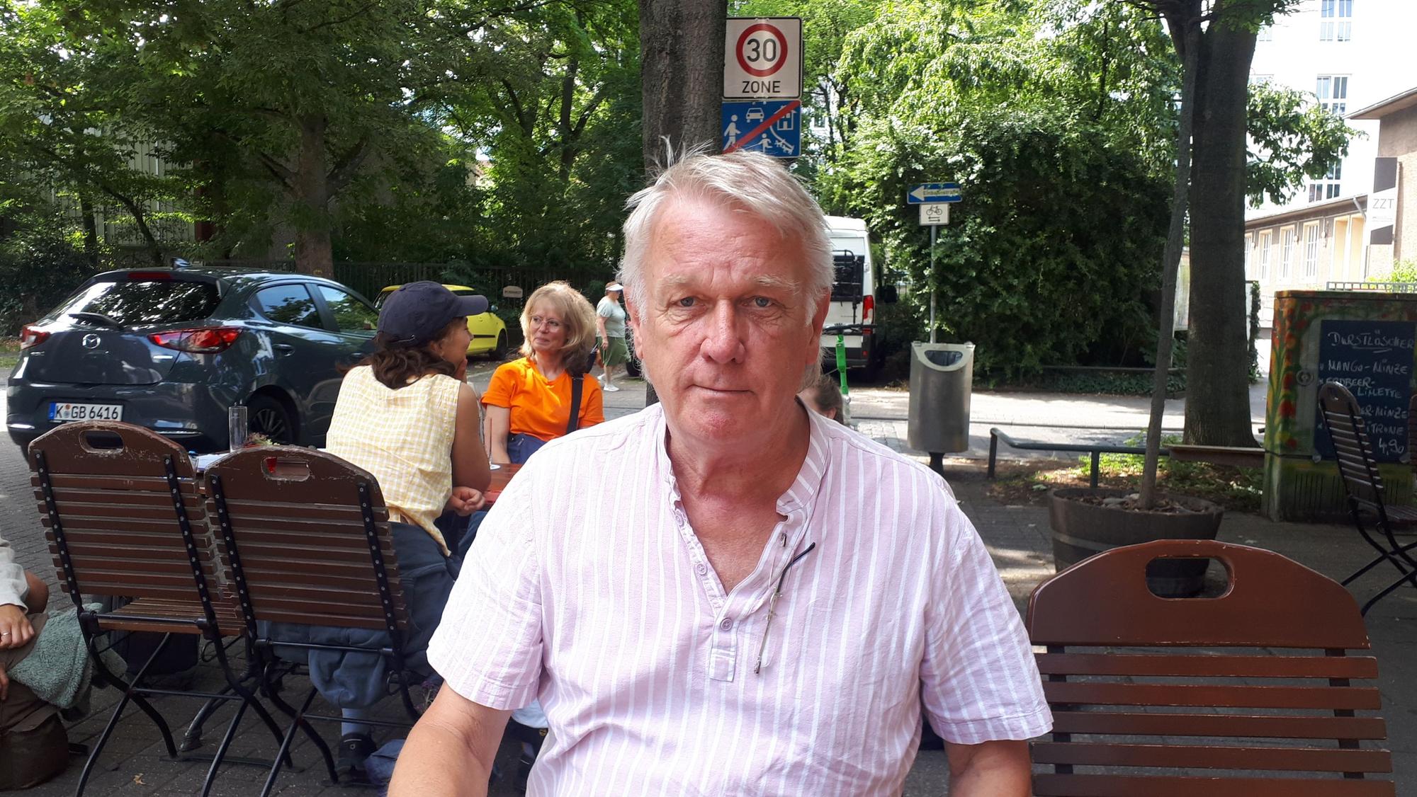 Ein Mann mit weißem Haar und hellem Hemd sitzt an einem Tisch im Außenbereich eines Cafés.