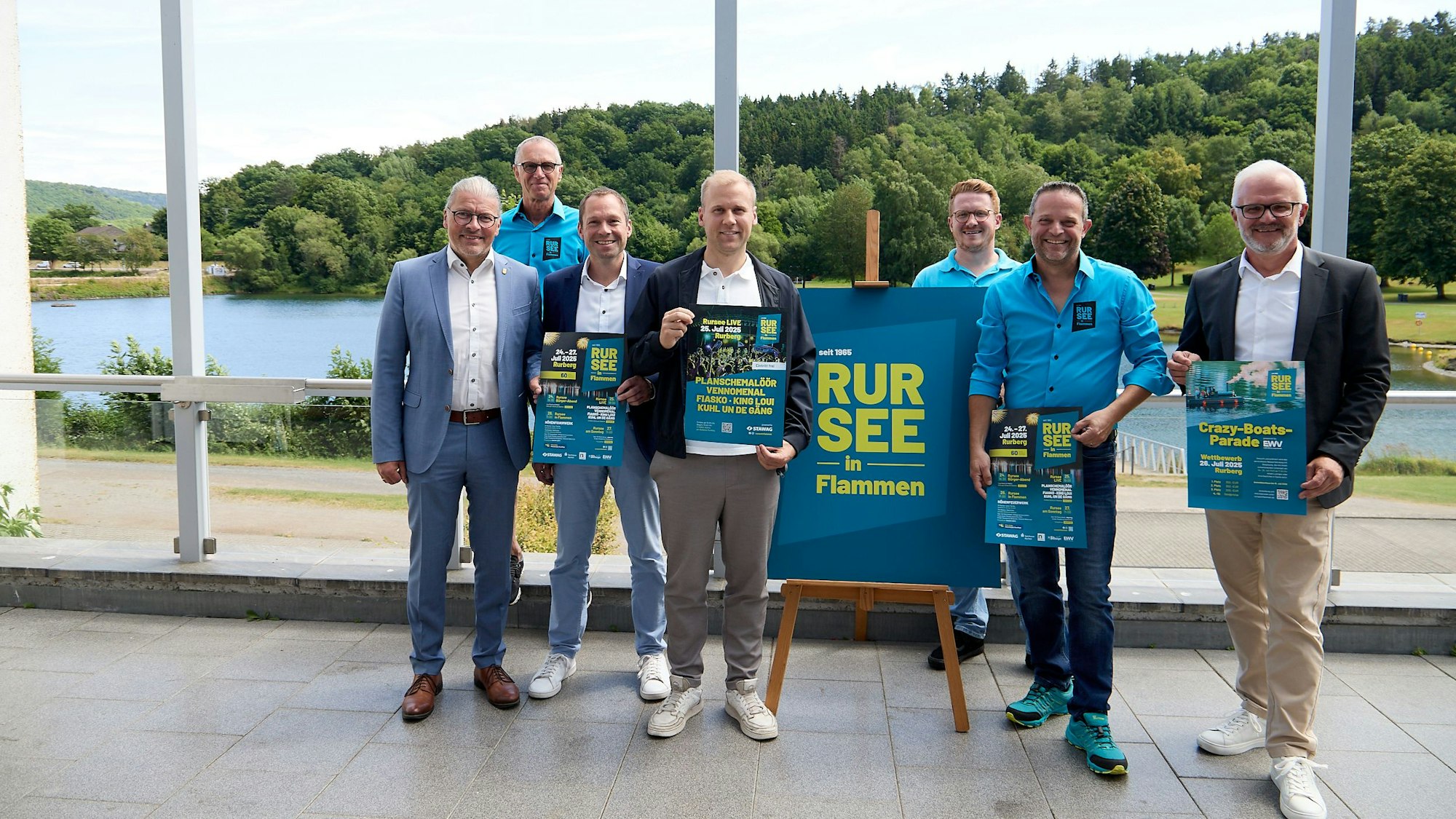 Sieben Männer zeigen Plakate vom „Rursee in Flammen“. Der Rursee befindet sich mit einem Wald im Hintergrund.