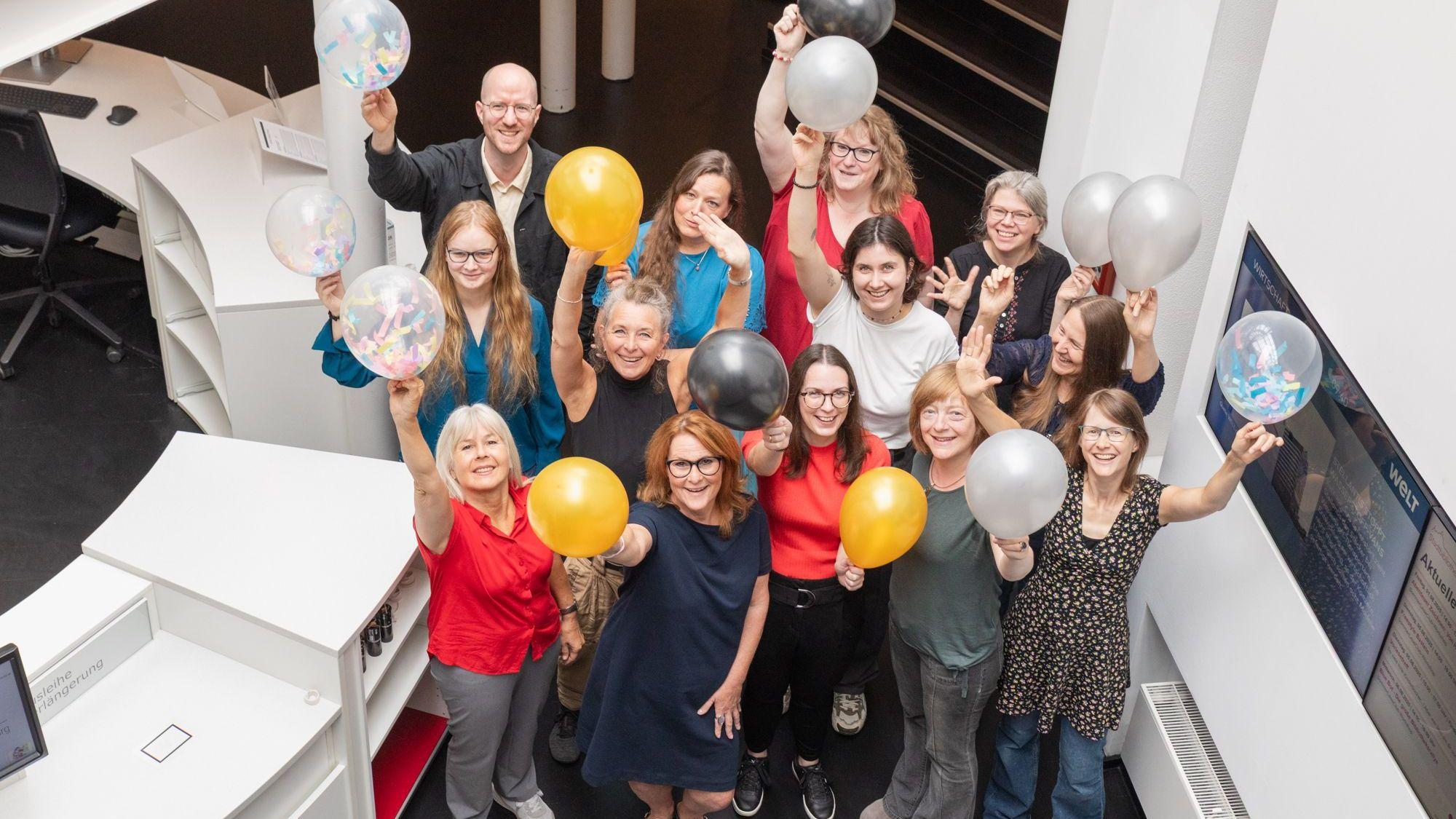 Frauen und ein Mann halten Luftballons in die Höhe. Sie freuen sich.