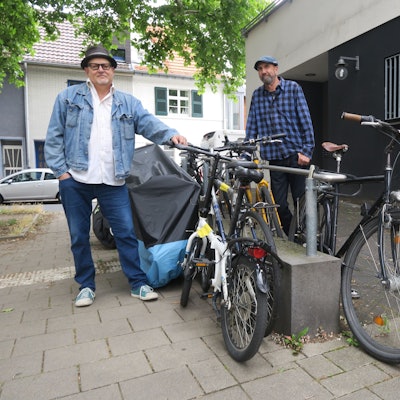Zwei Männer stehen neben abgestellten Fahrrädern an der Rampe zum Eingang einer ehemaligen Postfiliale.