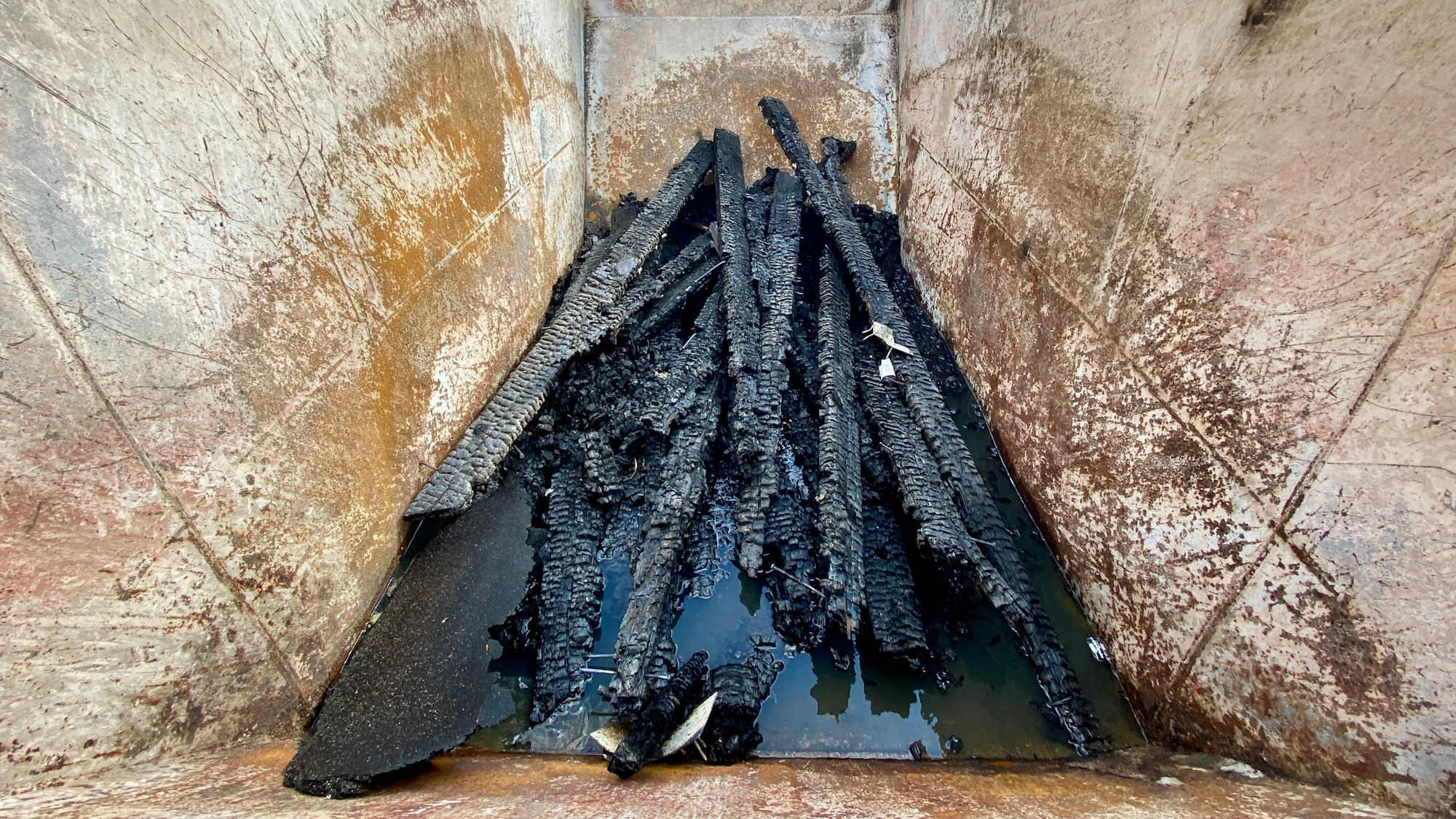 Im Inneren eines Schuttcontainers sieht man schwarz verkokelte Holzstücke.