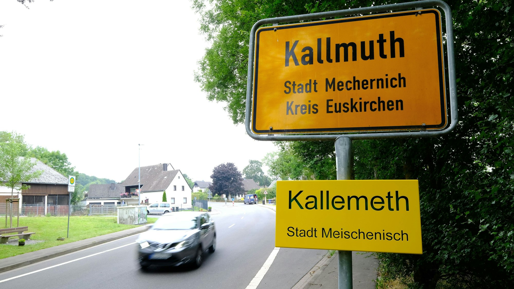 Ortstafel des Mechernicher Stadtteils Kallmuth mit Zusatzschild des Ortsnamens in Eifeler Mundart („Kallemeth, Stadt Meischenisch“). Auf der Dorfstraße fährt ein Auto vorbei.
