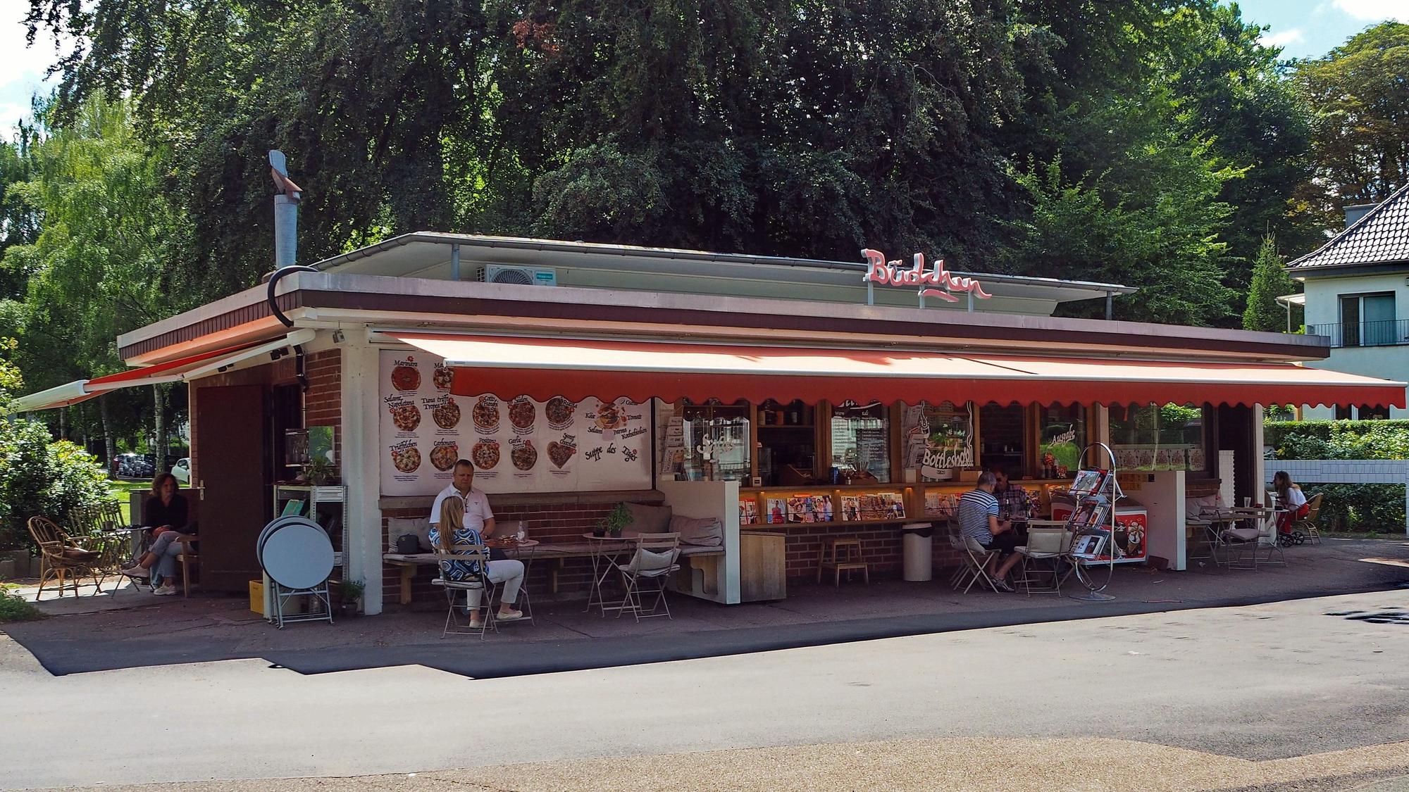 Der Kiosk an der Endhaltestelle der Buslinie 106 ist ein langgestrecktes Flachdach-Gebäude, auf dem Dach ist der Schriftzug „Büdchen“ angebracht.