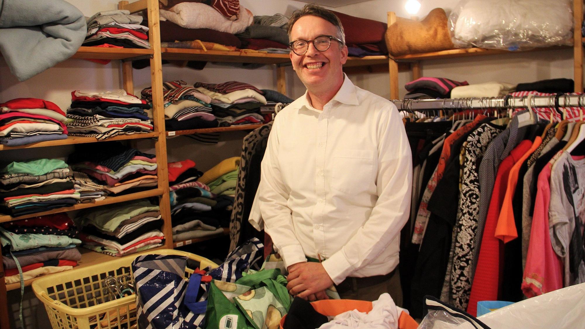 Ein Mann mit grauem Haar und Brille steht inmitten von Regalen und Kleiderstange mit Kleidung.