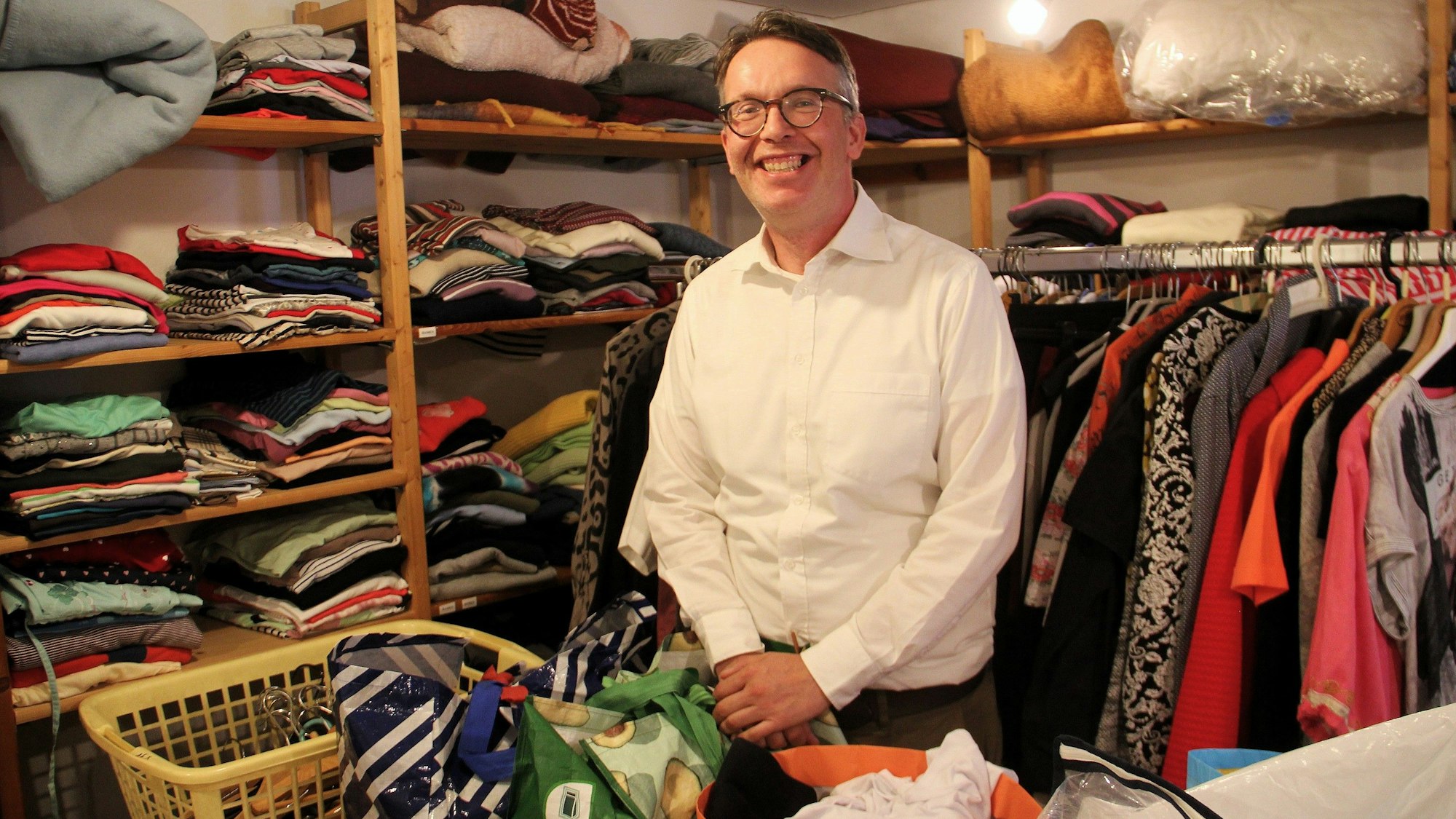 Ein Mann mit grauem Haar und Brille steht inmitten von Regalen und Kleiderstange mit Kleidung.