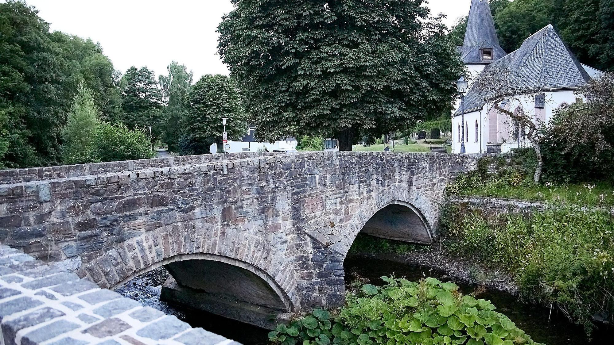 Eine Bogenbrücke führt in Hellenthal-Blumenthal über die Olef. Im Hintergrund ist eine Kirche zu sehen.