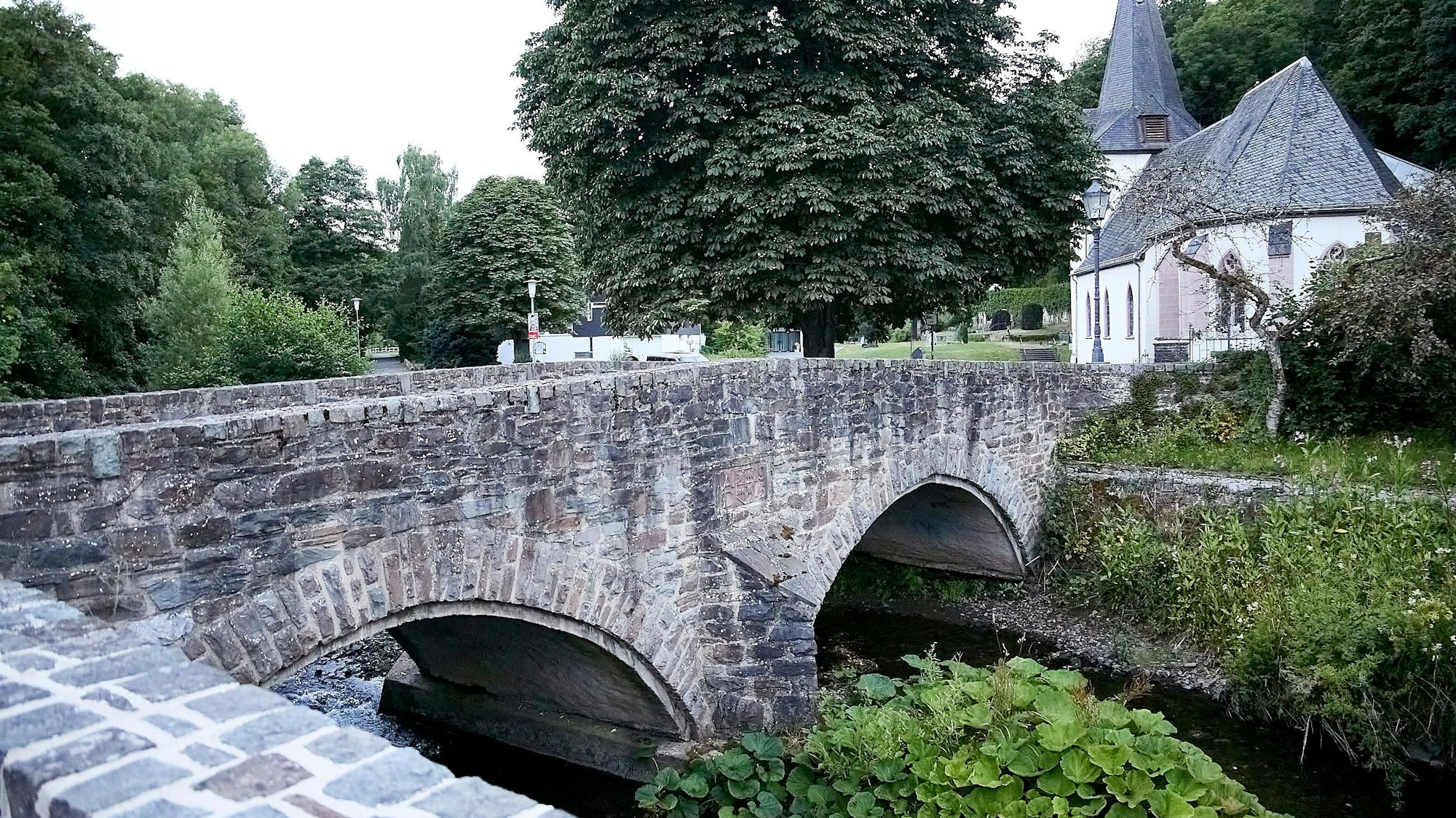 Eine Bogenbrücke führt in Hellenthal-Blumenthal über die Olef. Im Hintergrund ist eine Kirche zu sehen.