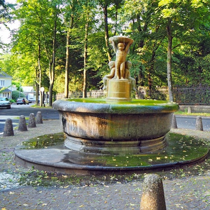 Ein runder Brunnen mit Kinderfiguren in der Mitte ist auf einem kleinen Platz umgeben von Pollern.