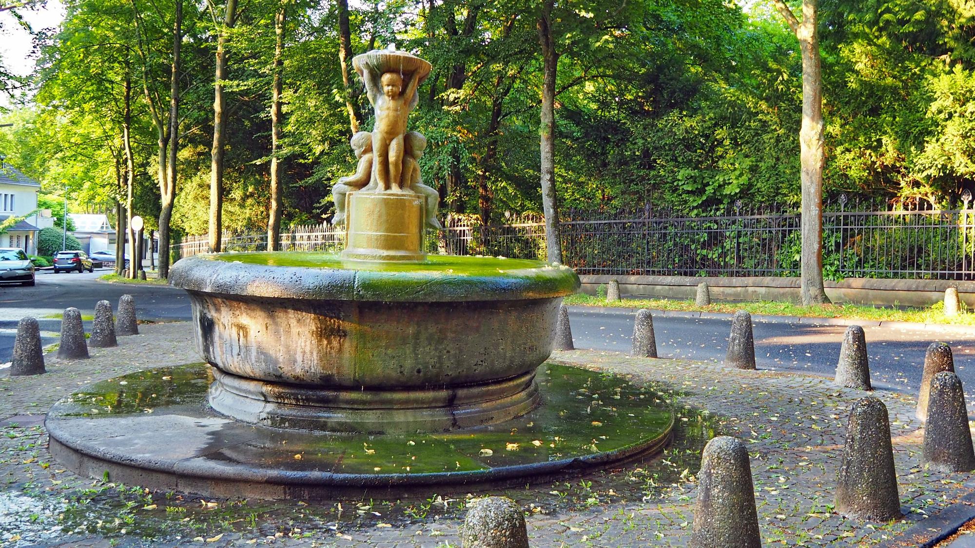 Ein runder Brunnen mit Kinderfiguren in der Mitte ist auf einem kleinen Platz umgeben von Pollern.
