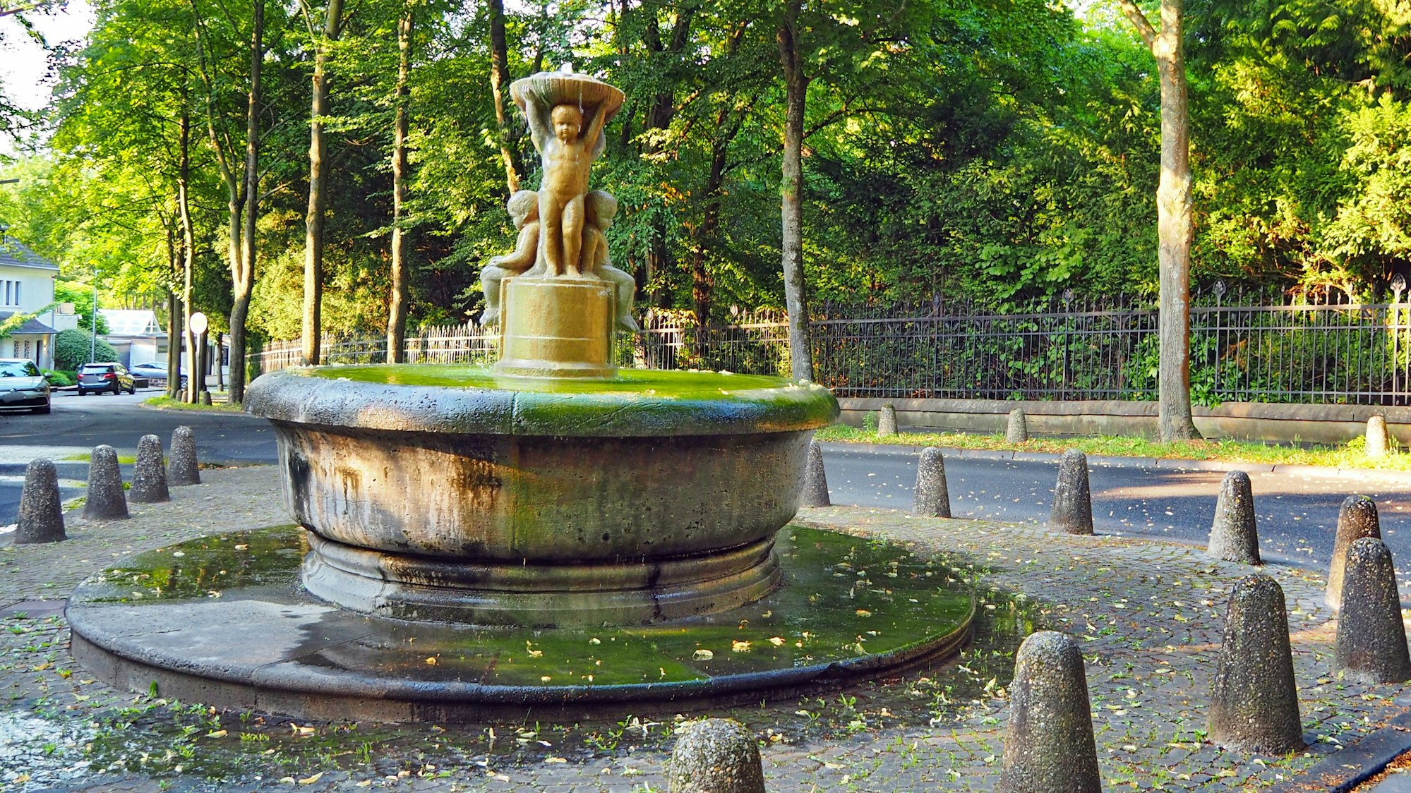Ein runder Brunnen mit Kinderfiguren in der Mitte ist auf einem kleinen Platz umgeben von Pollern.