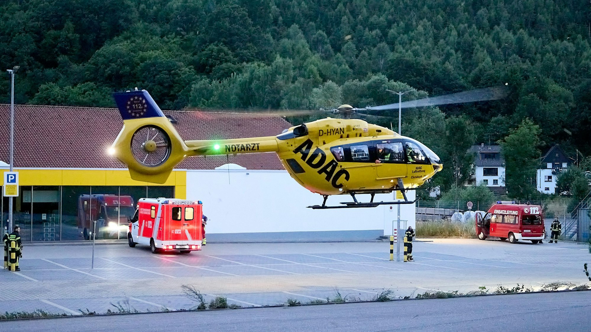 Ein gelber Rettungshubschrauber mit der Aufschrift ADAC landet auf dem Parkplatz des Netto-Marktes in Hellenthal. Darauf stehen ein Rettungswagen und der Einsatzleitwagen der Feuerwehr.