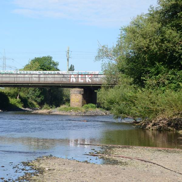 Eine Brücke über einen kleinen Fluss. Der Wasserstand ist niedrig, das Kiesufer ist breit.