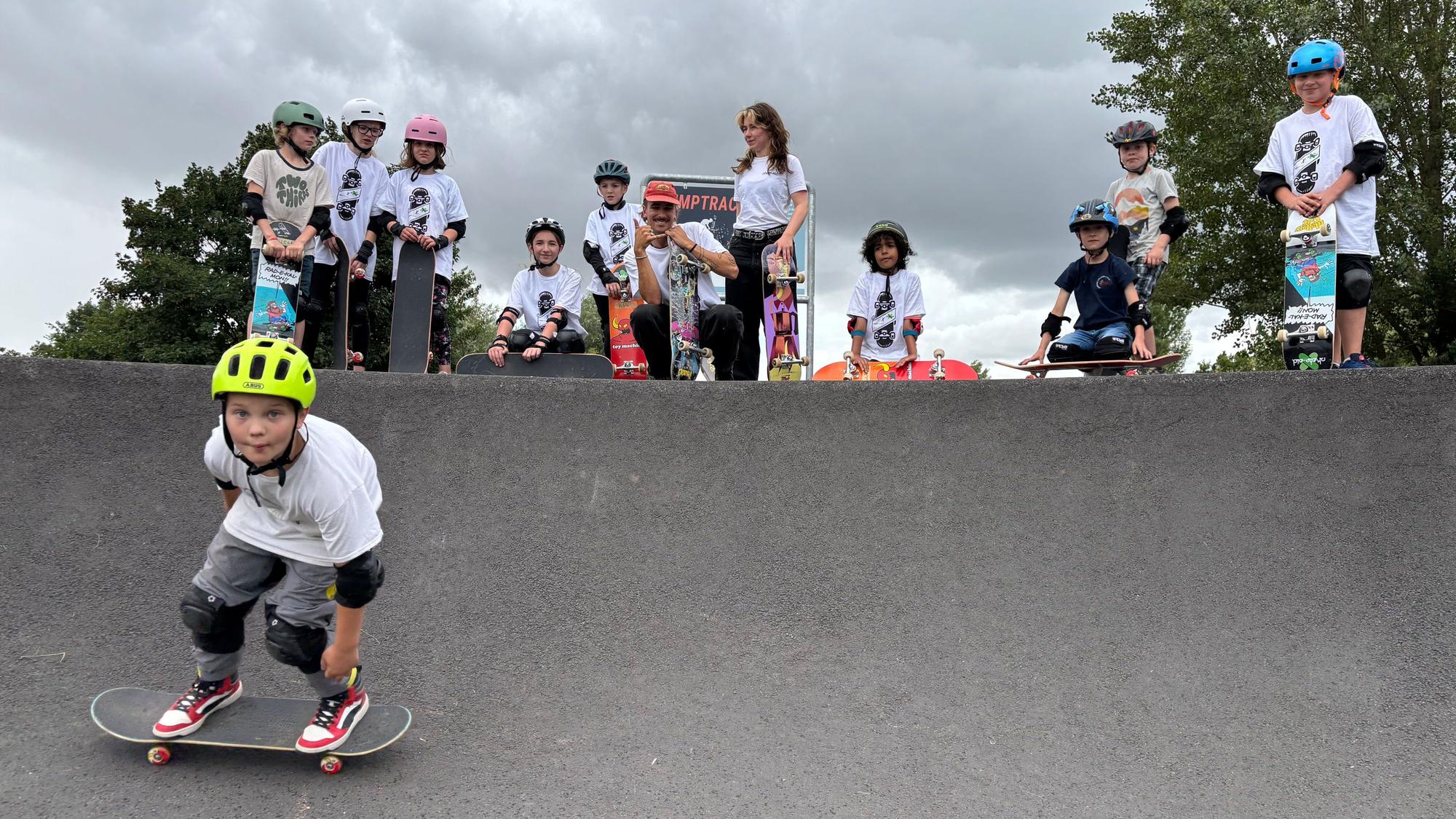 Ein Junge mit Helm und Schützern fährt auf einem Skateboard in eine Kurve des Weilerswister Pumptracks. Im Hintergrund sind andere Kinder, ein Betreuer und eine Betreuerin.