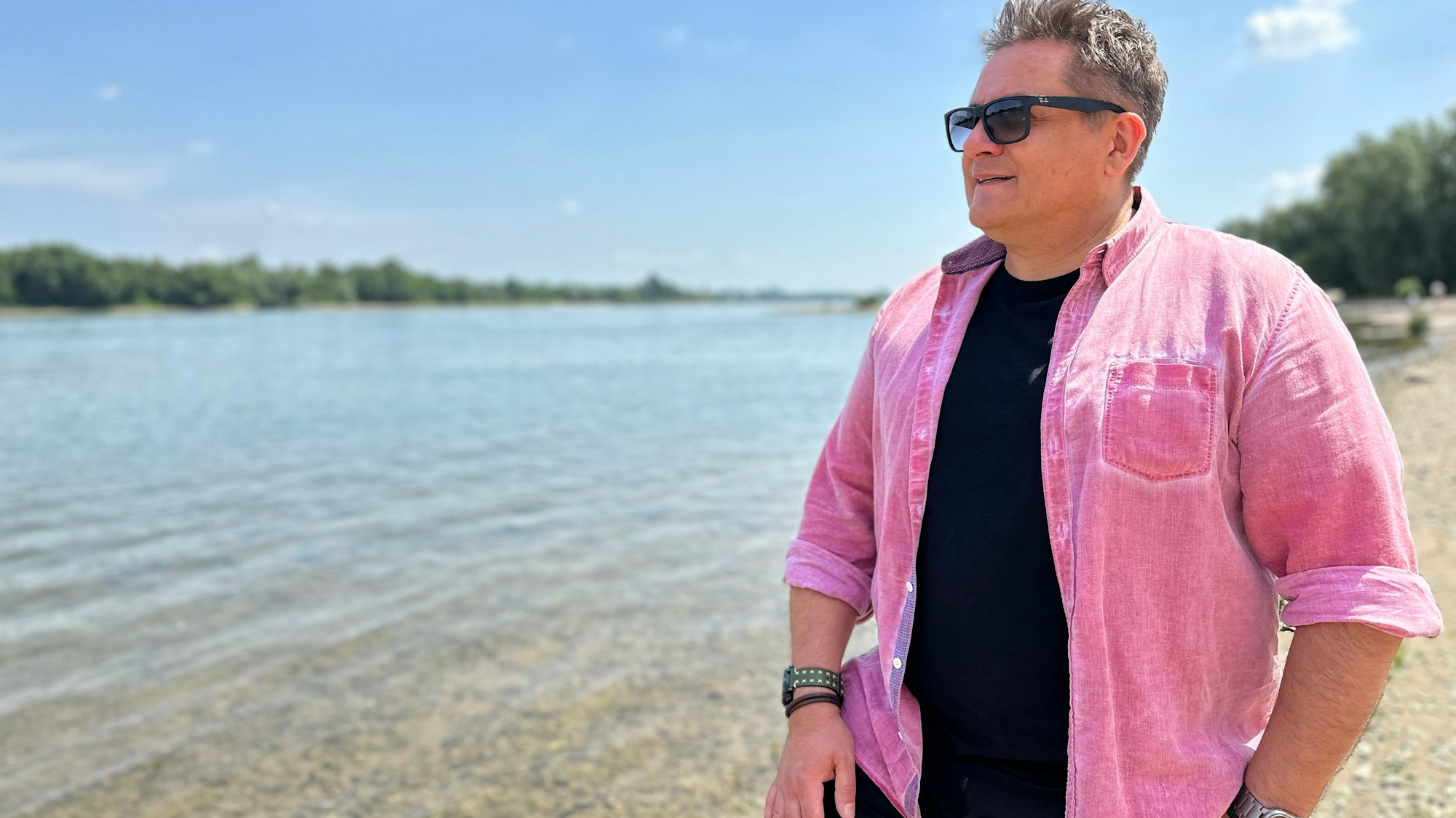 Ein Mann im rosa Hemd steht am Rheinufer und schaut auf den Fluss.