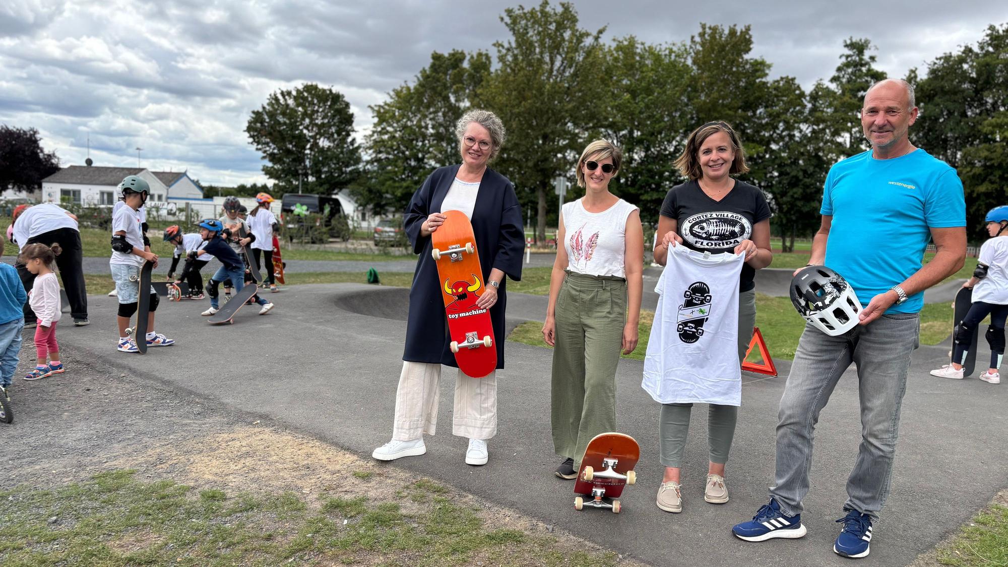 Anne Horst, Kerri Wetter, Stefanie Klein und Achim Diewald stehen auf dem Weilerswister Pumptrack. Die Bürgermeisterin hält ein Skateboard. Im Hintergrund stehen Kinder und Jugendliche mit Skateboards.