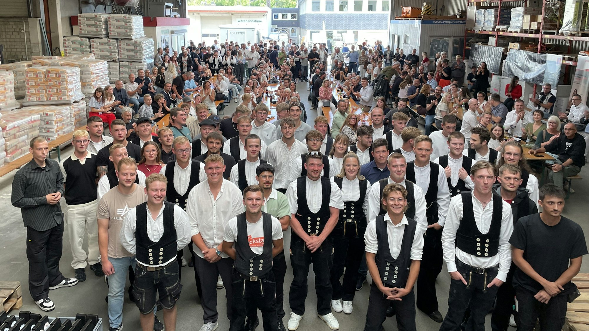 Alle 33 neuen Experten im Dachdecker- und Baugewerkshandwerk stehen für das Gruppenfoto parat. Dahinter haben sich Freunde und Familie auf Bierbänken versammelt.