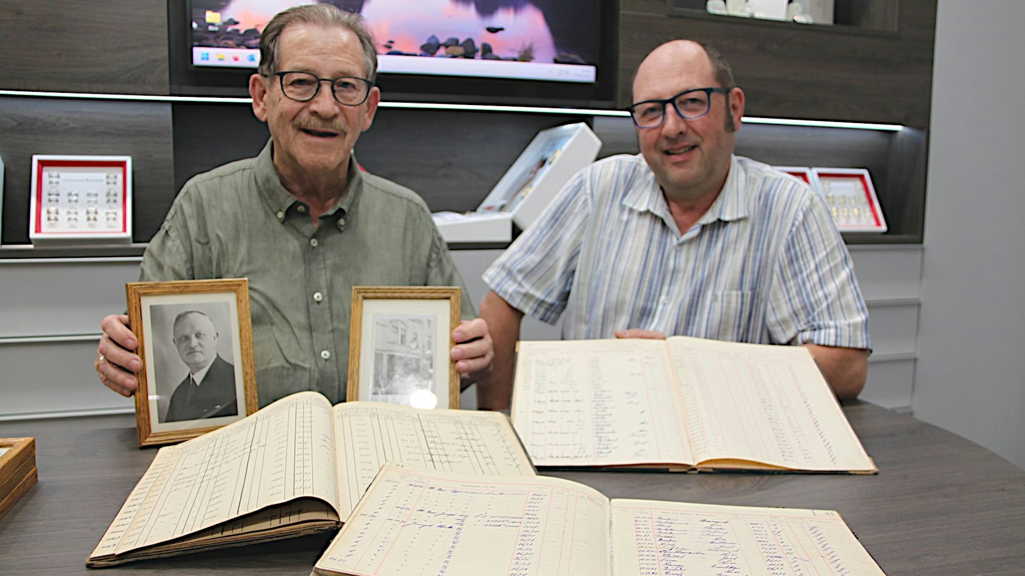 Walter und Michael Blumenthal, beide tragen Brille, sitzen vor aufgeschlagenen Büchern und halten ein Foto von Johann Joseph Blumenthal in die Kamera.