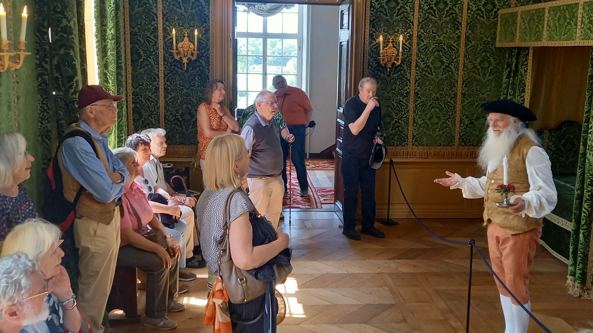 Eine Gruppe in einem hoheitlichen Schlafzimmer, es spricht ein Mann mit Bart und Kerze in der Hand.