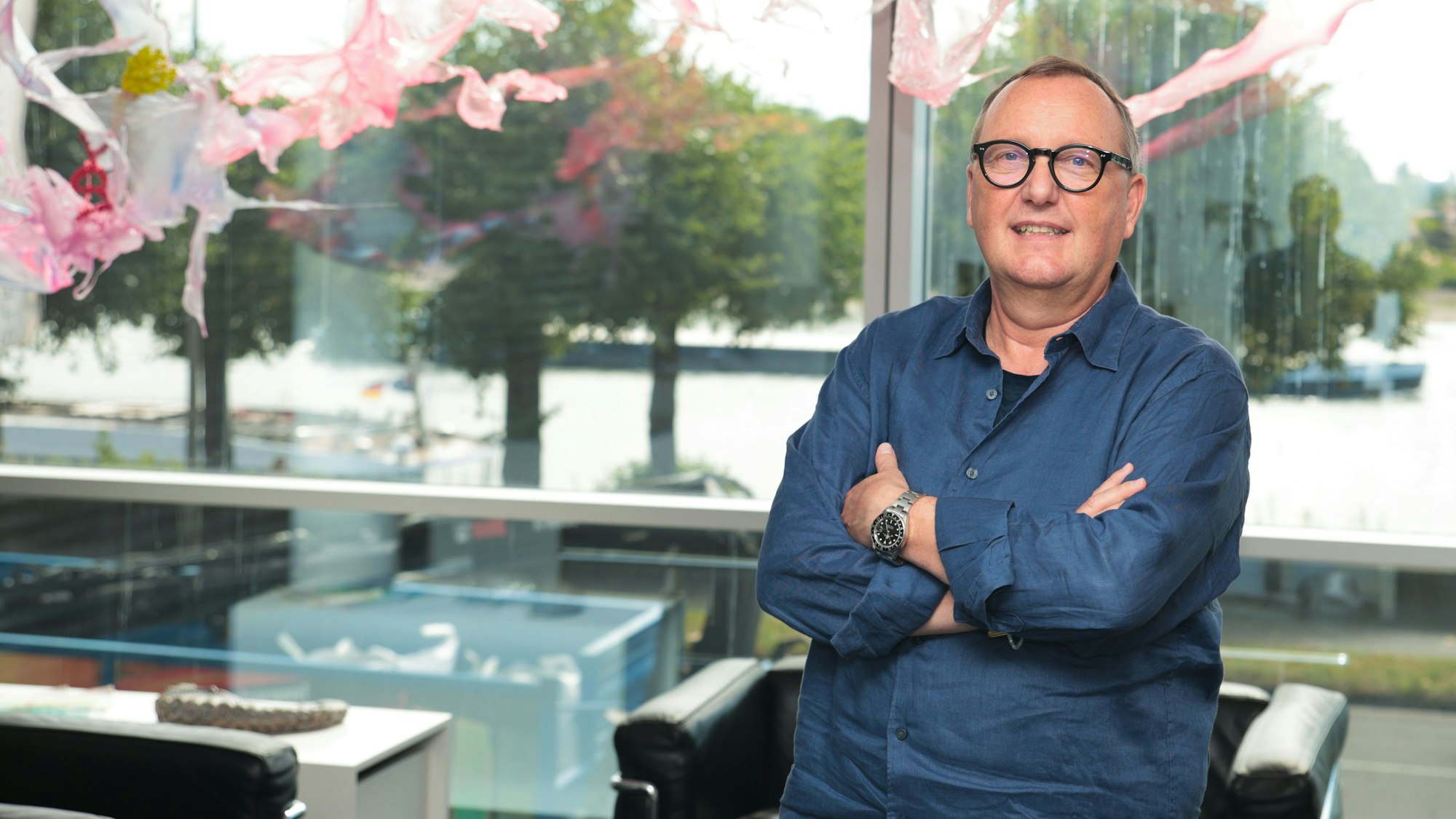 Ein Mann mit blauem Hemd und Brille steht vor einer Glasfront, hinter ihm fließt der Rhein.