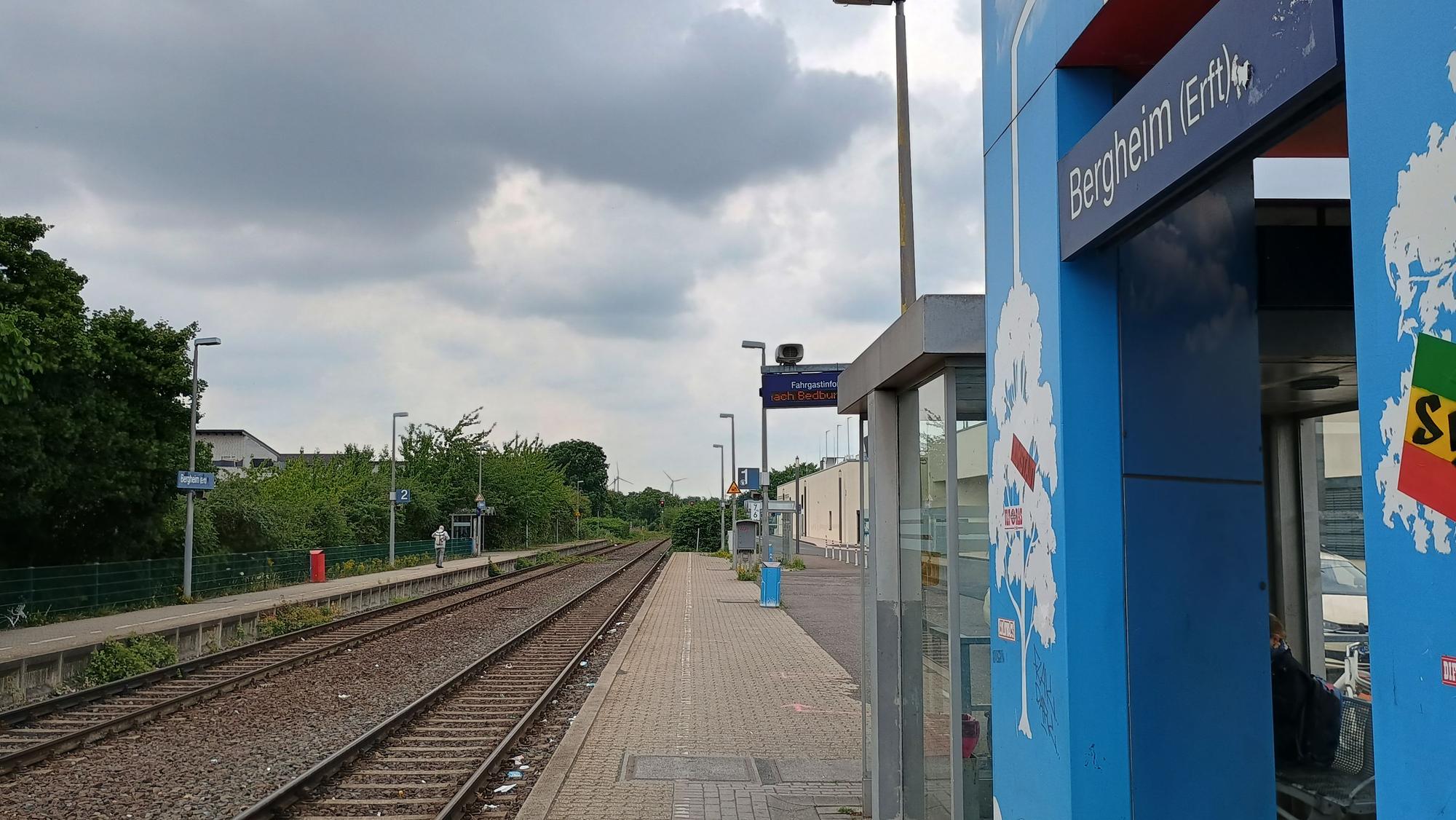 Das Bild zeigt die Bahngleise und daneben den Bahnsteig. Auf einem blauen Schild steht: „Bergheim (Erft)“