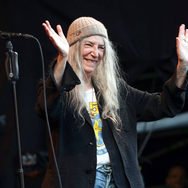 Patti Smith lächelt und winkt von der Bühne vor dem Kölner Dom. Sie trägt einen Beanie und ein weites schwarzes Jackett über einem T-Shirt.