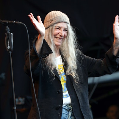Patti Smith lächelt und winkt von der Bühne vor dem Kölner Dom. Sie trägt einen Beanie und ein weites schwarzes Jackett über einem T-Shirt.