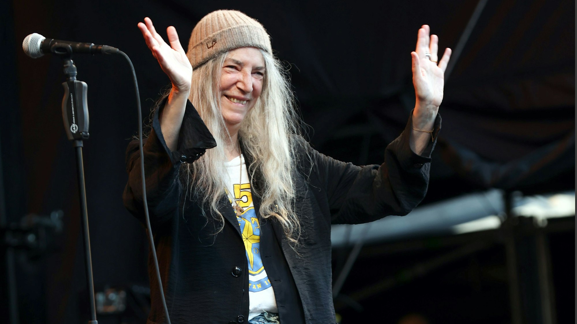Patti Smith lächelt und winkt von der Bühne vor dem Kölner Dom. Sie trägt einen Beanie und ein weites schwarzes Jackett über einem T-Shirt.