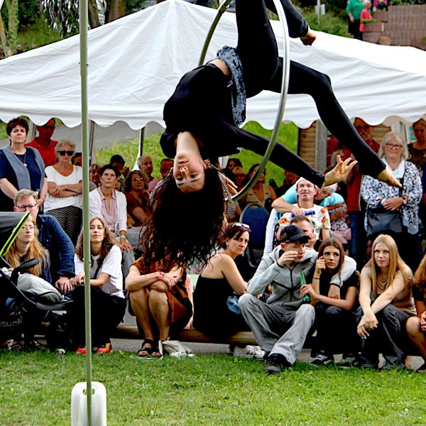 Die schwarz gekleidete Artistin hängt kopfüber in einem Stahlreifen, der mit Bändern an einer Stange befestigt ist. Im Hintergrund sind viele Zuschauer zu sehen.
