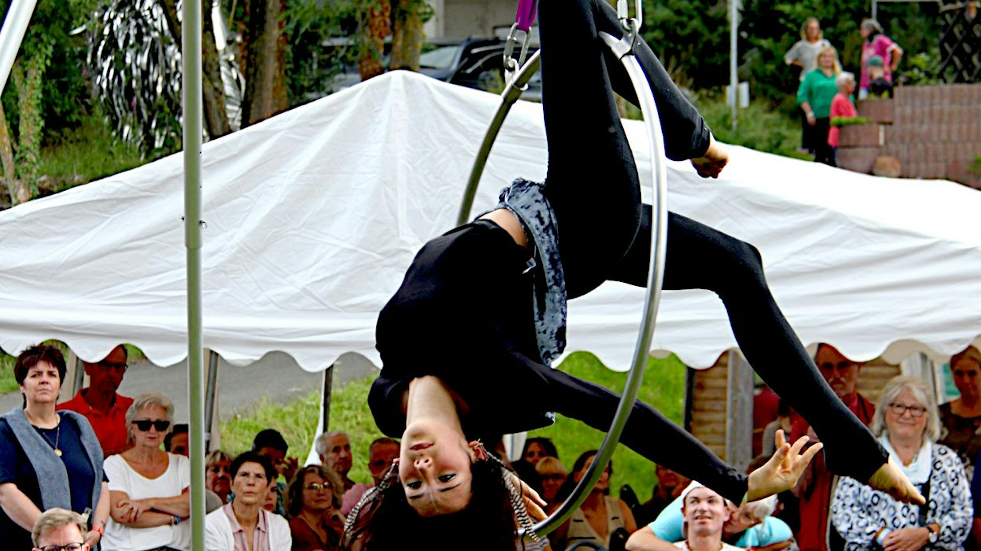 Die schwarz gekleidete Artistin hängt kopfüber in einem Stahlreifen, der mit Bändern an einer Stange befestigt ist. Im Hintergrund sind viele Zuschauer zu sehen.
