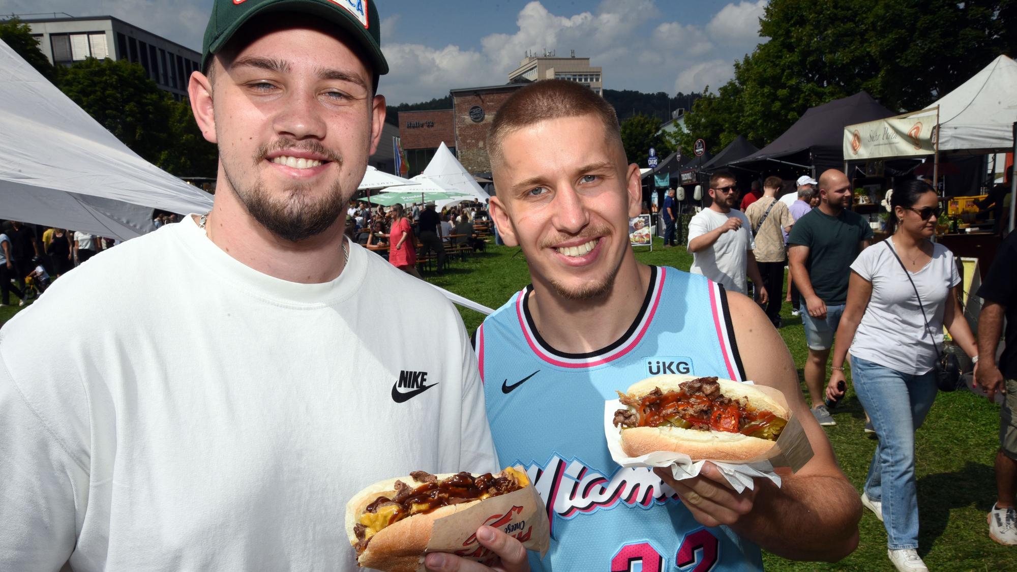 Zwei Männer halten ihr Essen grinsend in die Kamera.
