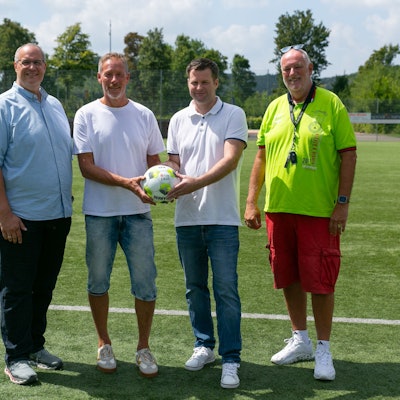 Vier Männer stehen nebeneinander. Die beiden in der Mitte halten gemeinsam einen Ball in den Händen.