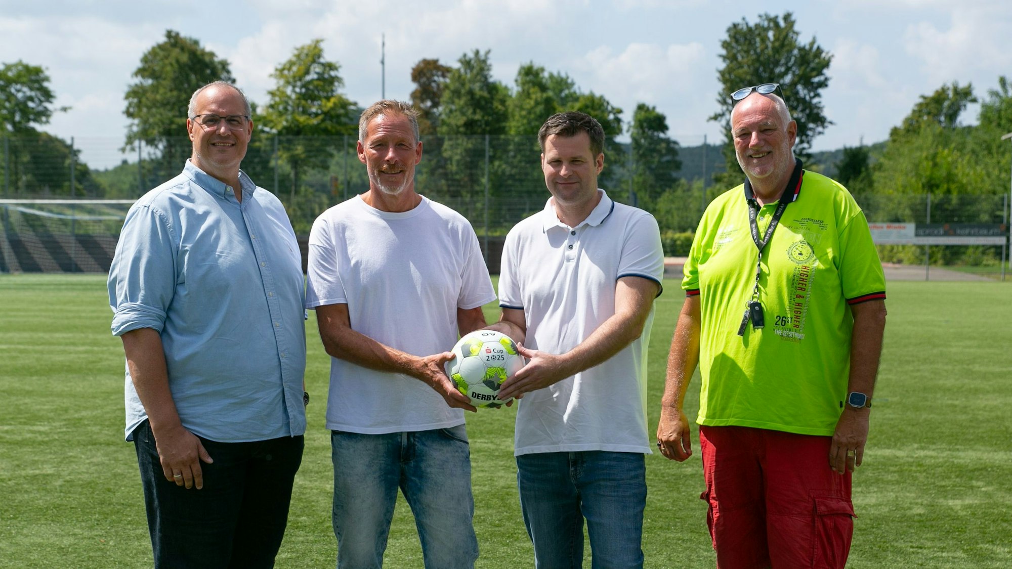 Vier Männer stehen nebeneinander. Die beiden in der Mitte halten gemeinsam einen Ball in den Händen.