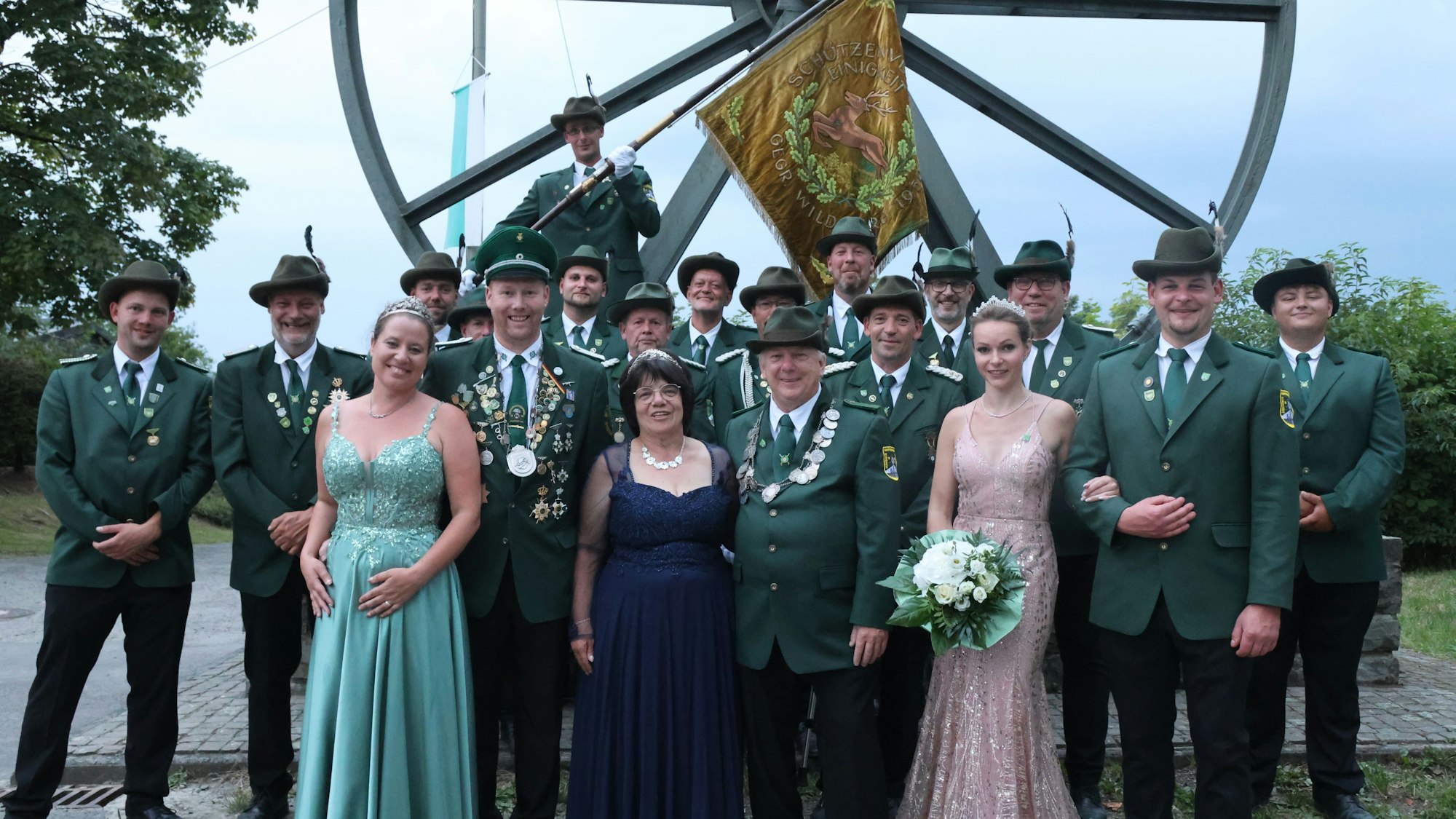 17 Männer in grünen Uniformen und drei Frauen in Kleider stehen vor einem Riesenrad.