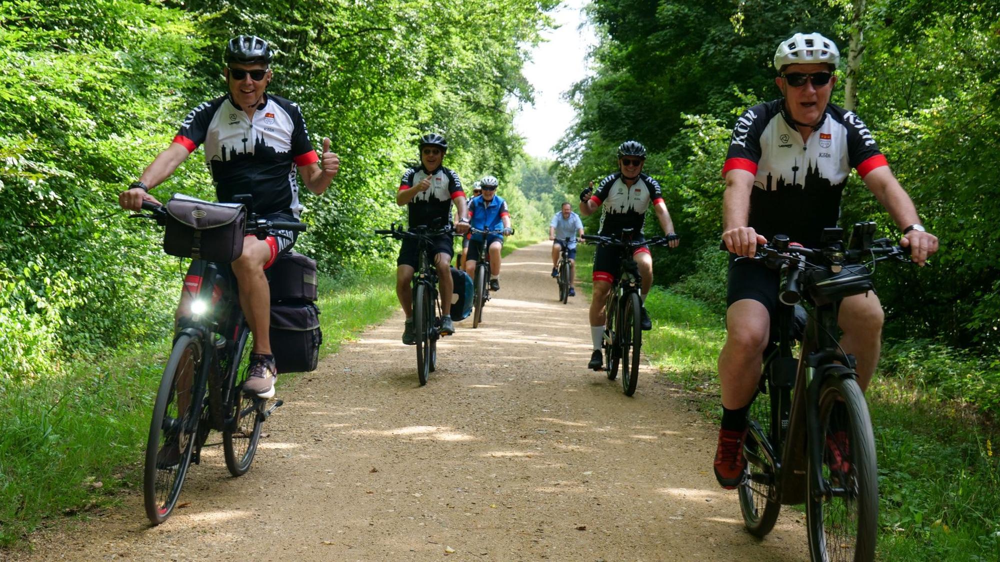 Die Fahrradfahrer tragen Trikots, auf denen Kölns Silhouette zu sehen ist. Sie fahren durch einen bewaldeten Weg.