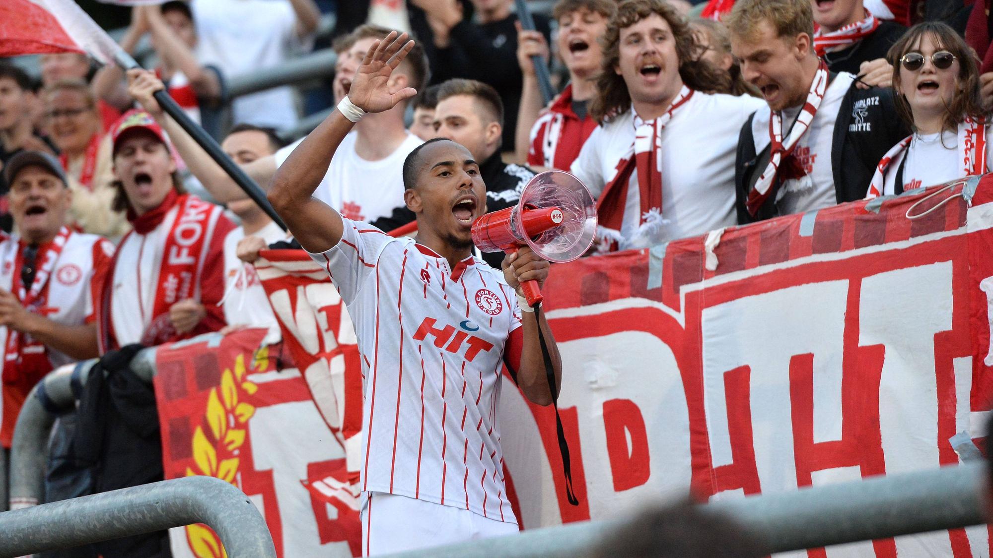 Robin Afamefuna ist im Trikot und mit Megafon im Stadion vor Publikum zu sehen.