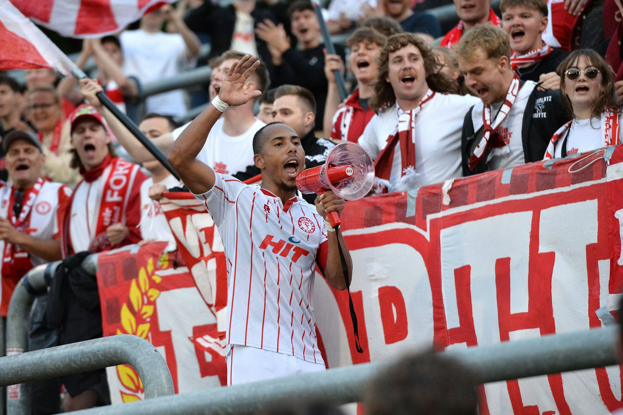 Robin Afamefuna ist im Trikot und mit Megafon im Stadion vor Publikum zu sehen.
