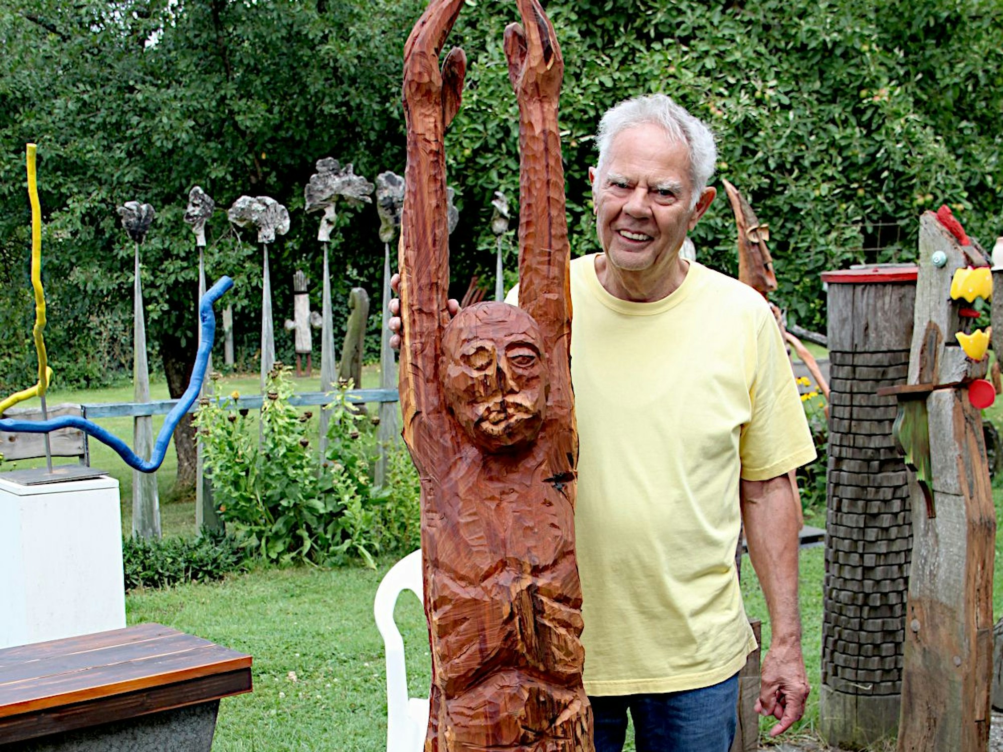 Ein Künstler steht neben einer Holzfigur, die die Arme hochstreckt.