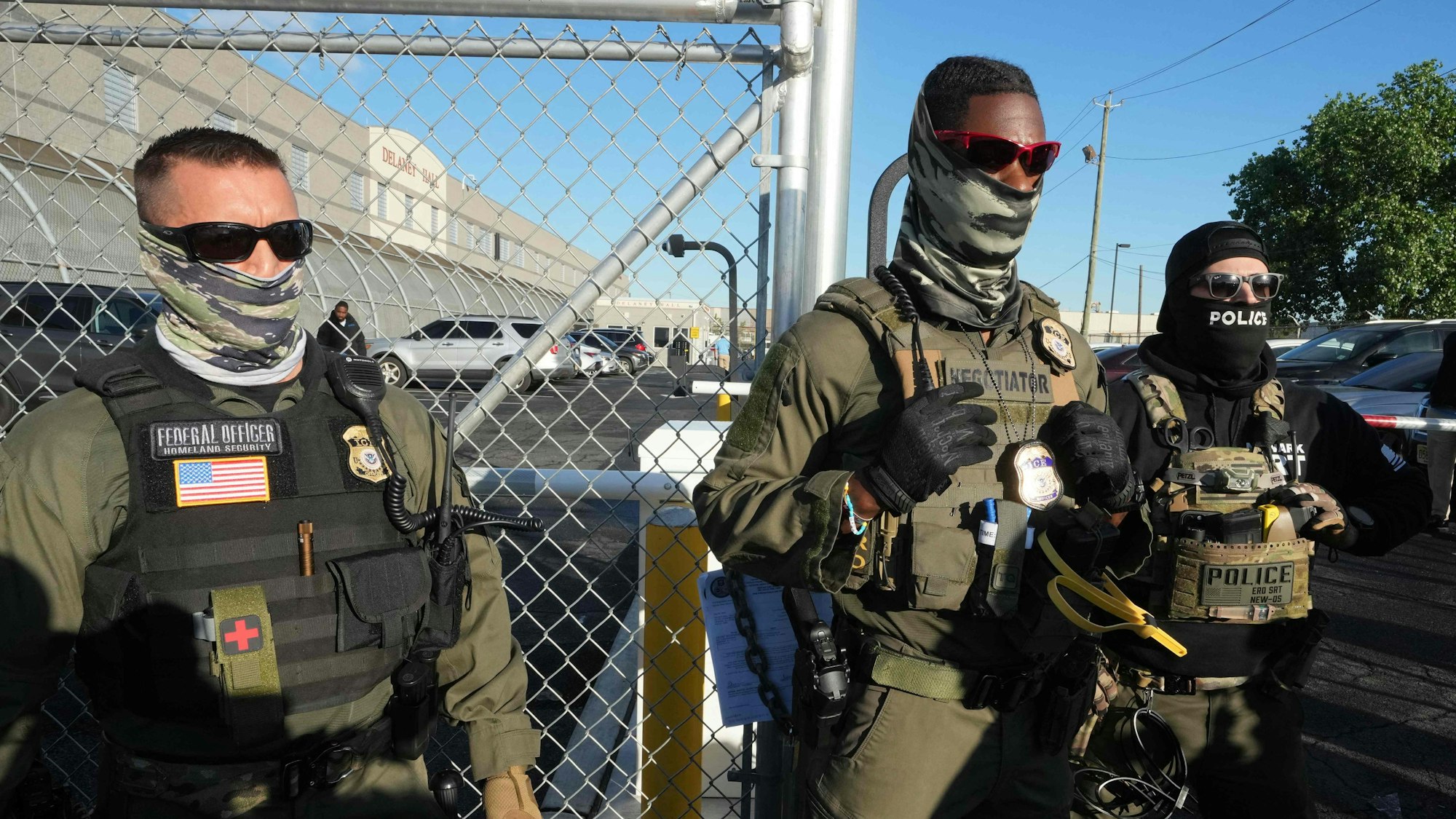 ICE-Beamte stehen neben einem Maschendrahtzaun.