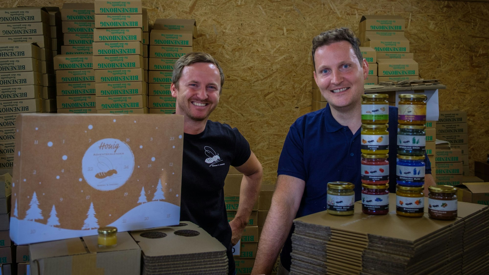 Zwei Männer stehen in einem Lagerraum mit vielen Verpackungskartons. Rechts im Bild sind verschiedene Honigsorten aufeinander gestapelt.