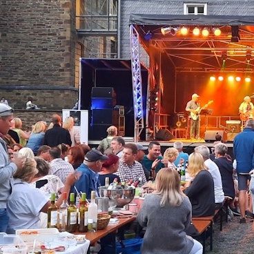 Eine Open-Air-Bühne. Davor sitzen Menschen auf Bierzeltbänken an Tischen.